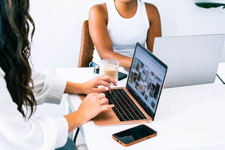 Two women working on laptops at a white desk, one browsing Pinterest-style content, with a phone and coffee nearby, representing evergreen blogs, content planning and digital marketing collaboration.