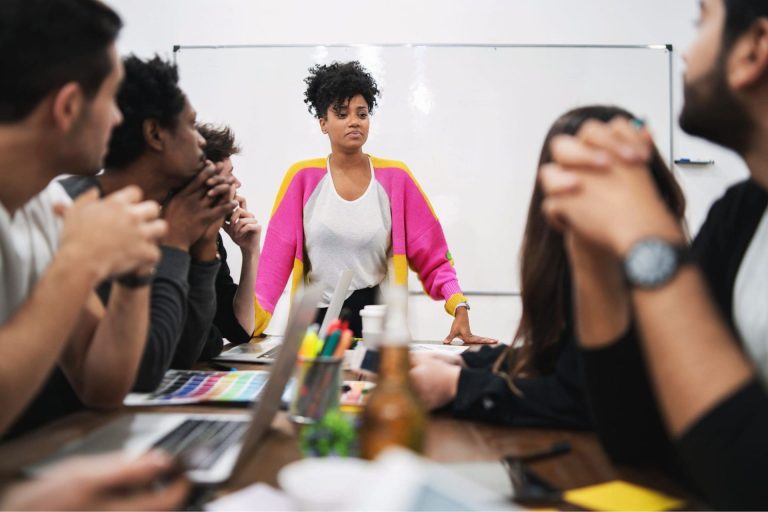 Team meeting with a woman leading a marketing strategy discussion at a whiteboard, surrounded by laptops and planning tools.