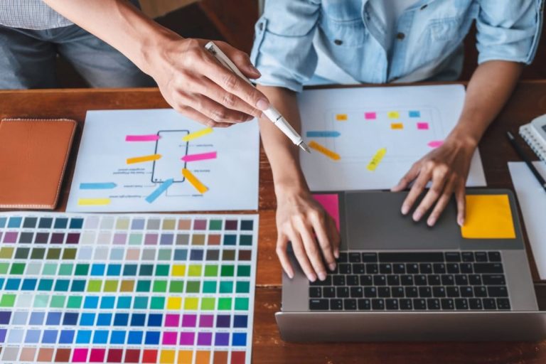 Designer planning website layout with sticky notes and color swatches, working on a laptop to create a visually appealing and functional site with must have website features.