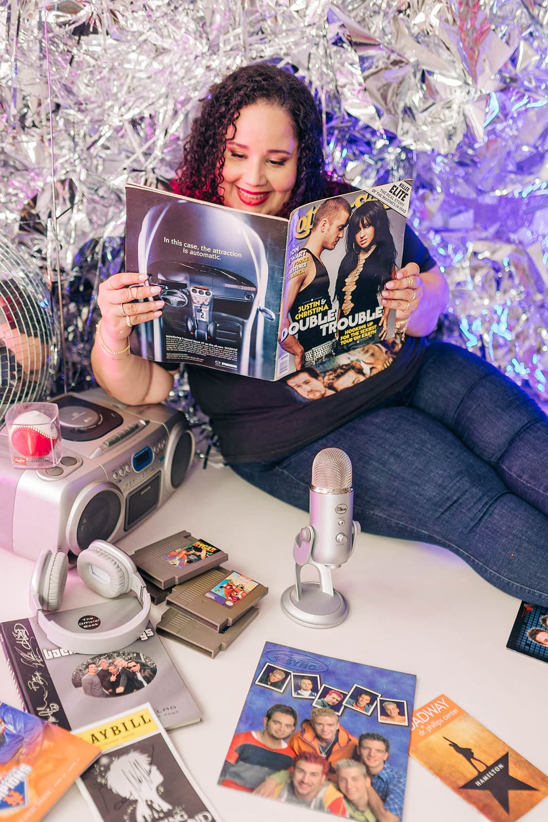 Kristine Gonzalez lounges against a backdrop of metallic streamers, smiling while reading a vintage Rolling Stone magazine featuring Justin Timberlake and Christina Aguilera. The table in front of her is filled with nostalgic items like a boombox, Nintendo cartridges, a silver microphone, ’90s and early 2000s memorabilia, and fan materials for Backstreet Boys, *NSYNC and Broadway shows.