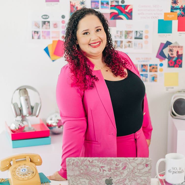 Kristine Gonzalez stands confidently at her desk in a bright, creative workspace, wearing a vibrant pink suit with a black top. Behind her is a colorful mood board filled with branding materials, swatches and inspiration images, highlighting her bold, design-forward aesthetic.