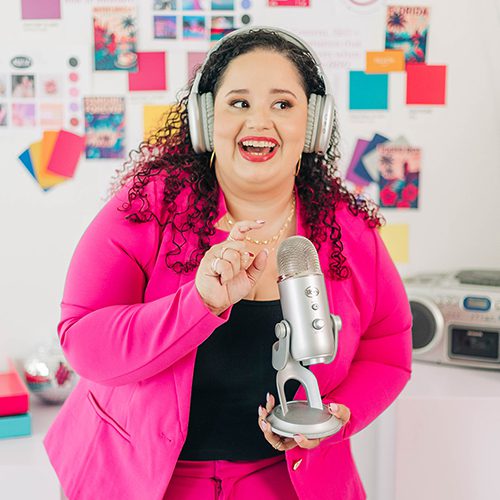 Kristine Gonzalez wears large headphones and smiles while speaking into a silver microphone, standing in a vibrant, design-filled office with a mood board behind her. Her hot pink suit pops against the creative, colorful workspace.