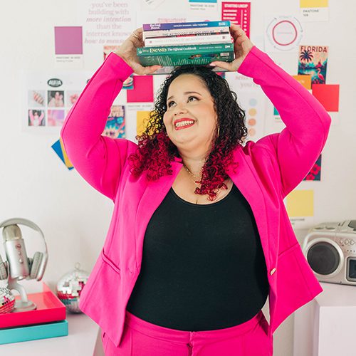 Kristine Gonzalez playfully balances a stack of marketing and business books on her head while beaming in a bright pink suit. The colorful background filled with swatches and design mockups reflects her fun and strategic brand personality.