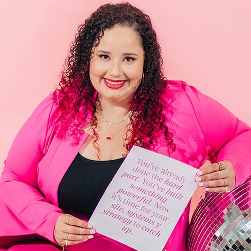 Latina entrepreneur in a vibrant pink suit seated beside a disco ball, holding a sign that reads: “You’ve already done the hard part. You’ve built something powerful. Now it’s time for your site, systems y strategy to catch up.”