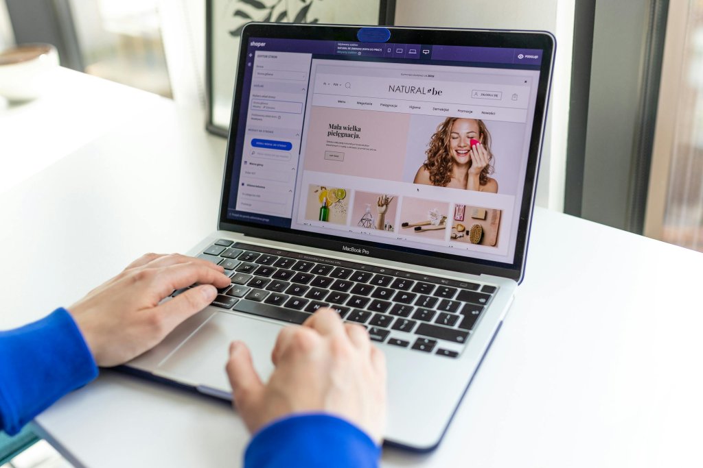 Hands type on a laptop displaying an ecommerce website homepage with product images and navigation menus visible on the screen. The image shows active website editing and content management, illustrating hands on work involved in maintaining and improving an online storefront.
