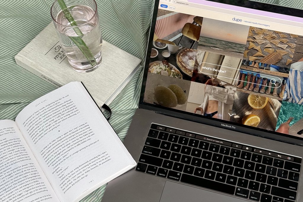 A laptop displaying a visual content grid sits beside an open book and a glass of water on a striped surface. The scene captures research and inspiration gathering to support clear website messaging.